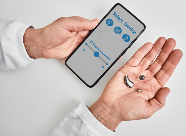 Doctor Audiologist Showing Smartphone App For Adjusting Hearing Aid Holding Smartphone In One Hand And Bte Hearing Aid In Other. Manage Hearing Aid Settings Via Smartphone